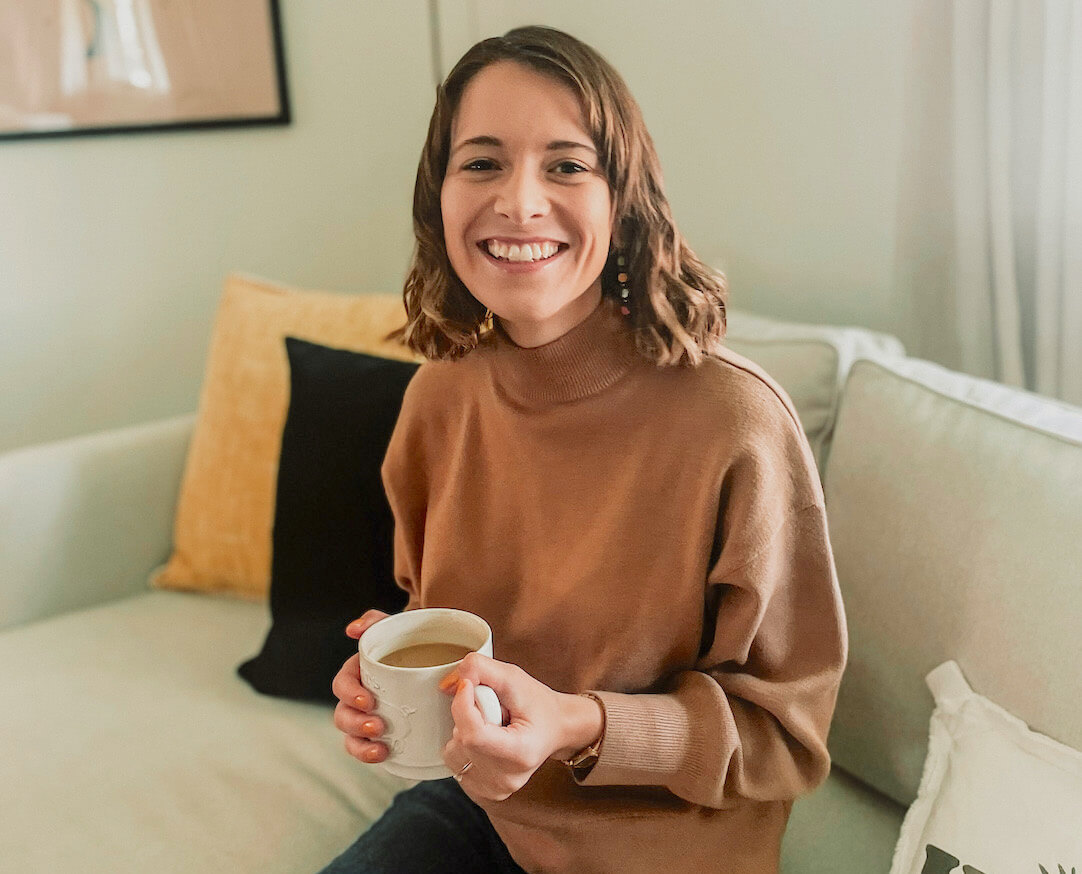 white woman with medium length brown wavy hair sits on a beige couch smiling and holding a mug of coffee