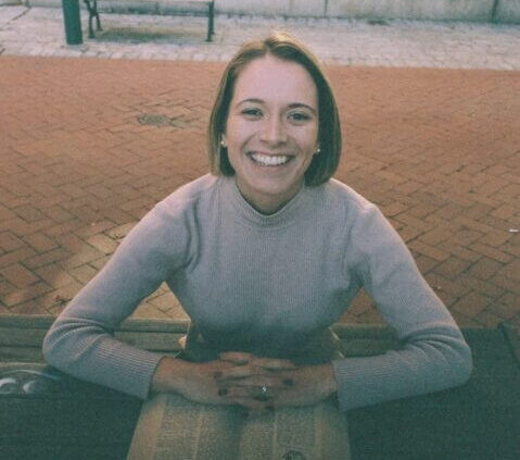 white woman with a short bob brunette haircut sits at a picnic table, her hands across an open book, smiling at the camera